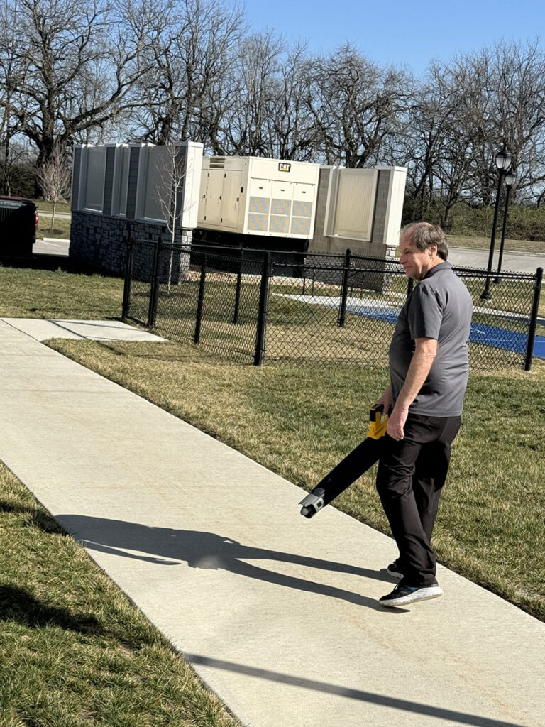 A community employee is using a leaf blower on a sunny day outside to clear the sidewalk.
