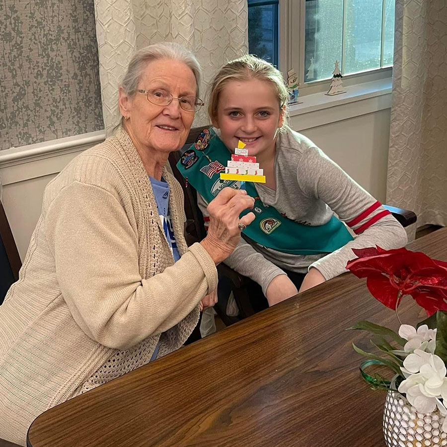 In this touching picture, an older woman and a Girl Scout show a paper birthday craft, brightening the day of memory care residents in the neighborhood.