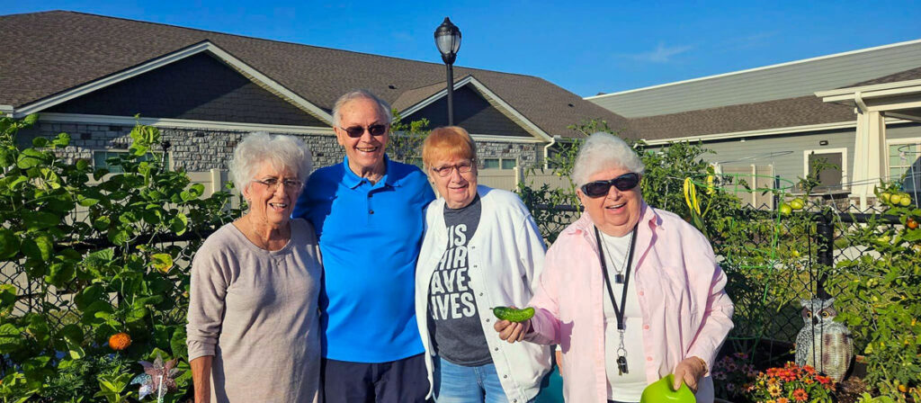 Group of senior living community residents smiling and standing together in a garden, with one resident holding a cucumber.