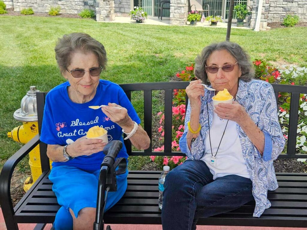 Two senior living community residents sitting on a bench outdoors, smiling and enjoying cups of frozen dessert.