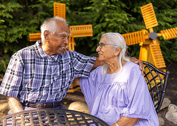 Two senior residents sit at a patio table, sharing a warm moment. The man has his arm around the woman, and they smile at each other with windmill decorations in the background.