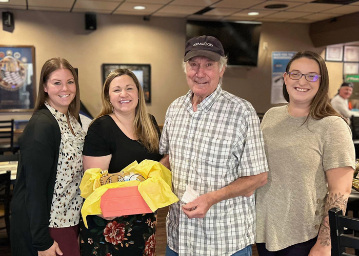 Four smiling individuals, including a senior resident, pose together indoors holding a vibrant gift box with yellow tissue paper, celebrating a special moment.