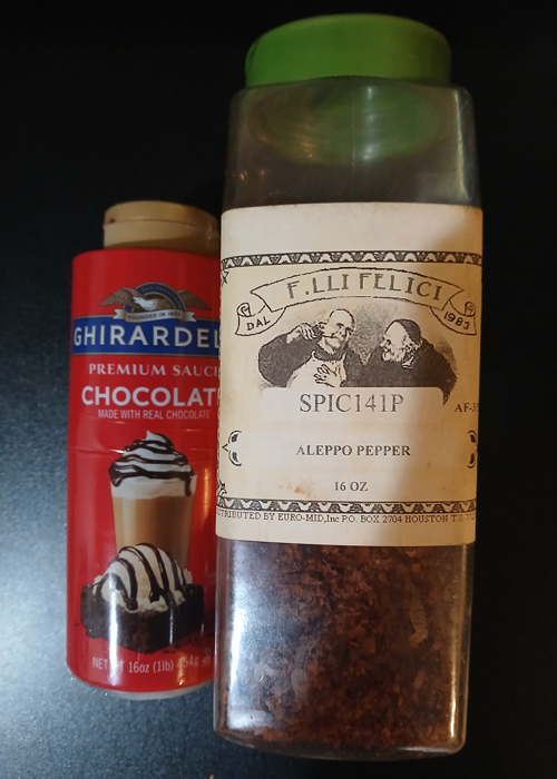 Ghirardelli chocolate sauce and a container of Aleppo pepper side by side on a dark counter, representing an unexpected yet flavorful ingredient pairing.