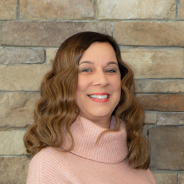 Jen Craigie, Business Office Director at The Princeton Senior Living, smiles warmly while wearing a pink turtleneck sweater in front of a tan stone wall.