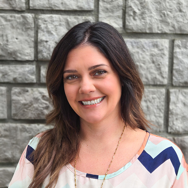 Colleen Elis, Memory Care Director at The Princeton Senior Living, smiles in front of a stone wall while wearing a pastel patterned blouse and gold necklace.