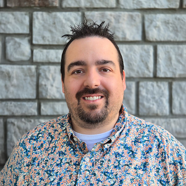 Justin Stewart, Senior Living Director for The Princeton Senior Living, smiles in front of a gray stone wall wearing a floral button-down shirt.