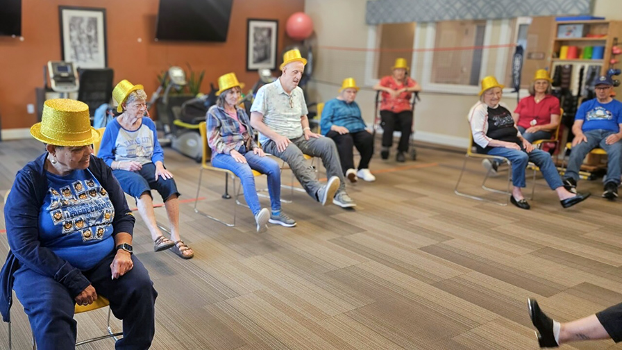 Group of senior residents wearing gold hats participate in a lively chair exercise session in the community wellness room.