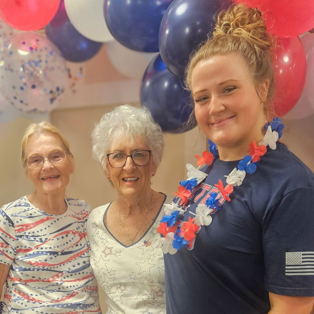 Two senior residents and a team member smile together at a patriotic celebration, surrounded by red, white, and blue balloons with festive attire and leis.
