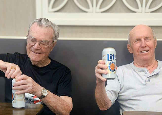 Two senior men sit together at a booth in the dining area of The Princeton, happily raising cans of beer.