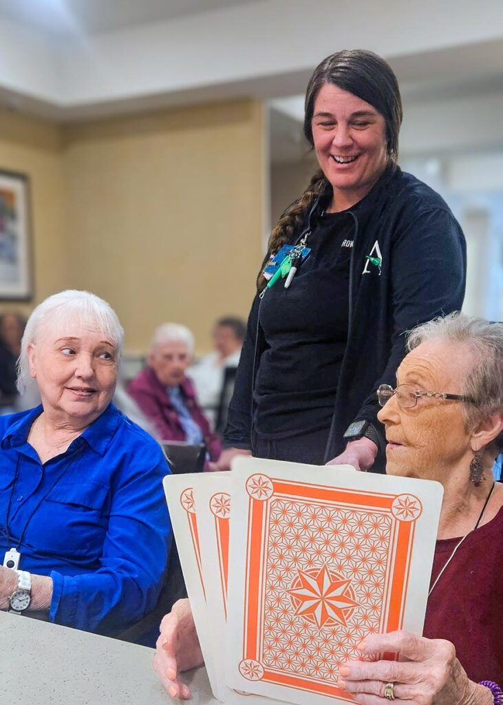 A team member looks on while two senior women play a game with jumbo cards.