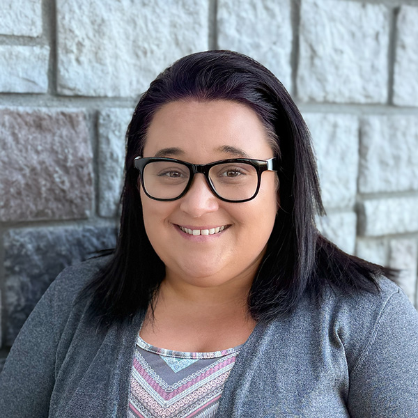 Jordan Sterns, Business Office Director at The Princeton Senior Living, smiling in glasses and a gray sweater while standing outdoors against a light stone wall.