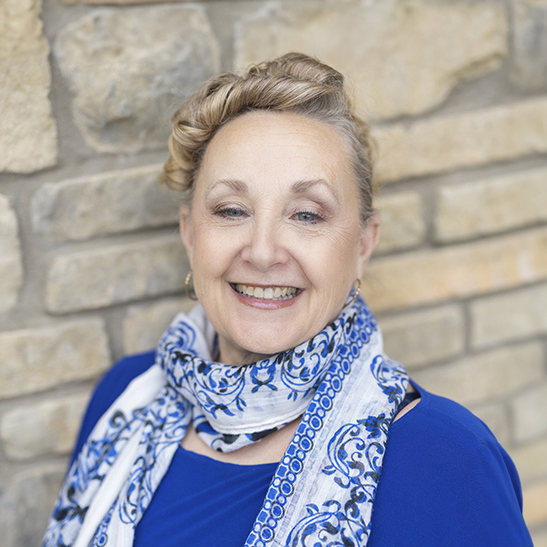Erin Winstead, Arrow Senior Living Regional Operations Specialist and Interim Executive Director at The Princeton Senior Living, smiling in a blue top and patterned scarf against a stone wall.