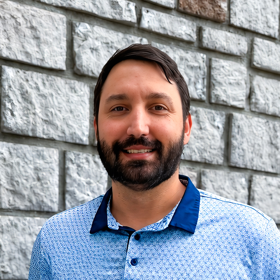 Daniel Tipton, Operations Coordinator at The Princeton Senior Living, smiling in a professional headshot with dark hair, a full beard, a blue patterned polo shirt, and a light gray stone wall background.