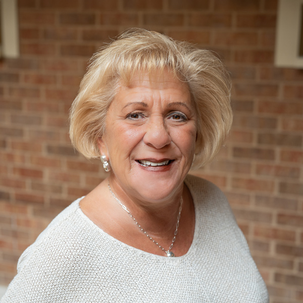 Melanie “Mel” McClain, Resident Services Director at The Princeton Senior Living, smiling in a professional outdoor headshot with short blonde hair, silver earrings, a light knit sweater, and a softly blurred brick background.