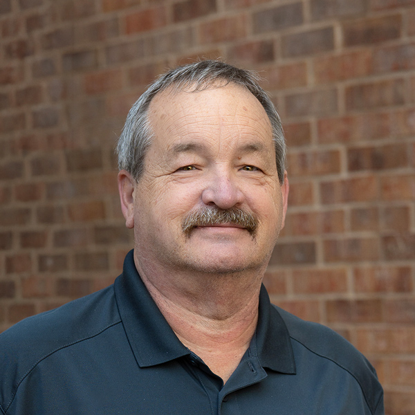 Tony Dudley, Plant Operations Director at The Princeton Senior Living, looking at the camera in a professional outdoor headshot with short gray hair, a mustache, a dark polo shirt, and a softly blurred brick background.