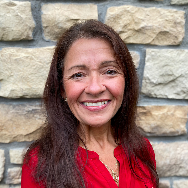 Andrea Groh, Senior Living Coordinator at The Parkway Senior Living supporting The Princeton Senior Living, smiling in a professional headshot with long dark hair, a red blouse, and a light stone wall background.