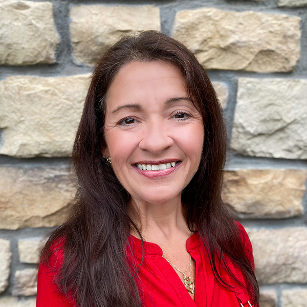 Andrea Groh, Senior Living Coordinator at The Parkway Senior Living supporting The Princeton Senior Living, smiling in a professional headshot with long dark hair, a red blouse, and a light stone wall background.