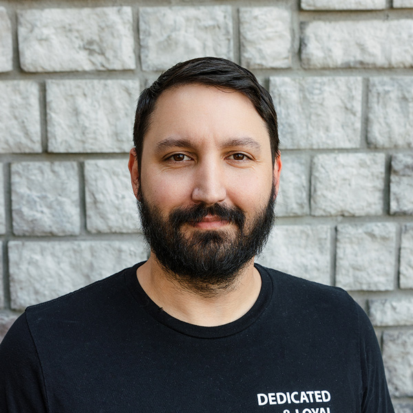 Daniel Tipton, Operations Coordinator at The Princeton Senior Living, looking at the camera in a professional headshot with short dark hair, a full beard, a black shirt, and a gray stone wall background.
