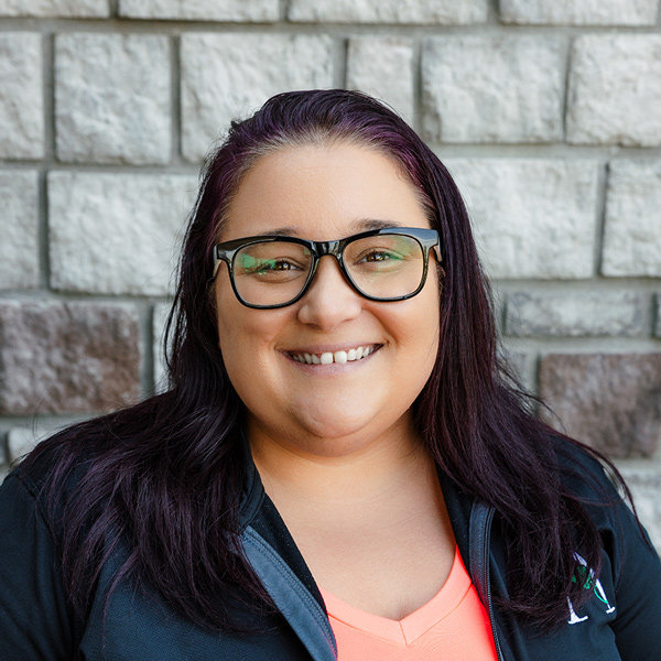 Jordan Sterns, Business Office Director at The Princeton Senior Living, smiling in a professional headshot with long dark purple hair, black glasses, a coral top, and a gray stone wall background.