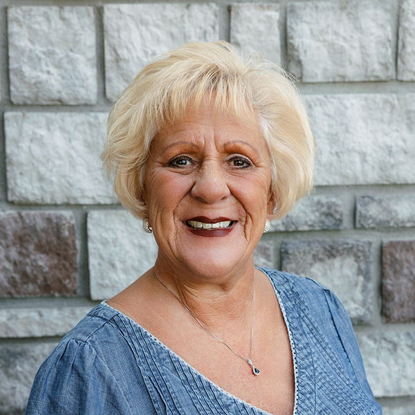Melanie “Mel” McClain, Resident Services Director at The Princeton Senior Living, smiling in a professional headshot with short blonde hair, silver earrings, a denim blue blouse, and a gray stone wall background.