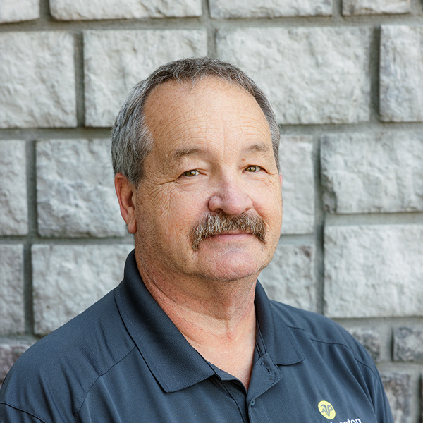Tony Dudley, Plant Operations Director at The Princeton Senior Living, looking at the camera in a professional headshot with short gray hair, a mustache, a dark polo shirt, and a gray stone wall background.
